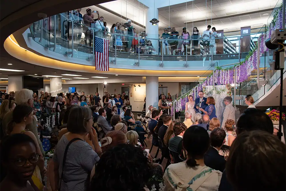 A large crowd gathers inside Arlington Public Library, watching a wedding ceremony beneath a staircase decorated with hanging flowers and an American flag.