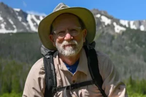 Mark Easter wearing a wide-brimmed hat and hiking gear with a mountainous landscape in the background.