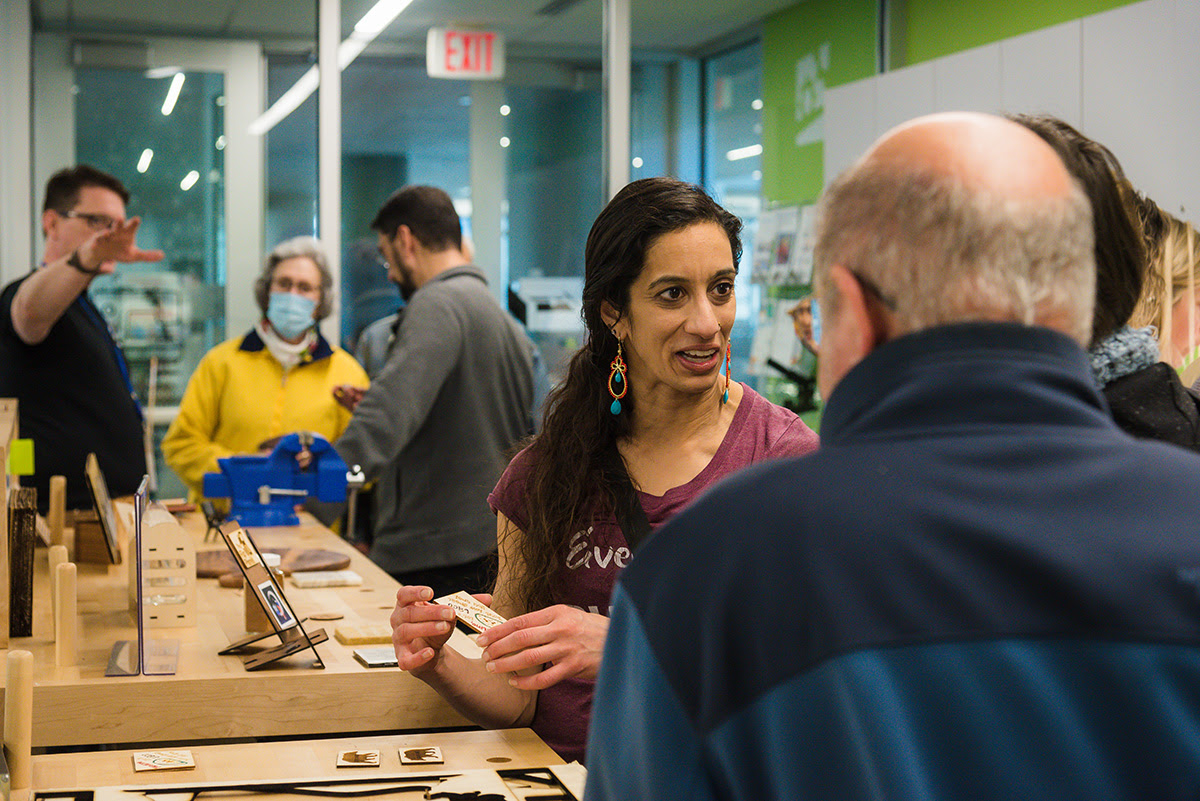 A woman in The Shop makerspace shows a laser engraved wood project to visitors as a staff member gives a tour in the background.