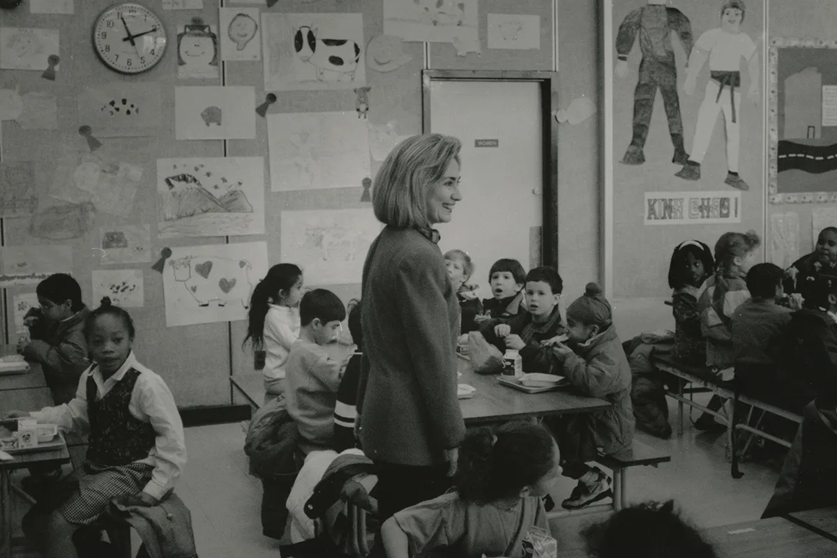 Hillary Clinton standing in the center of Longbranch Elementary School lunchroom talking to students eating lunch.