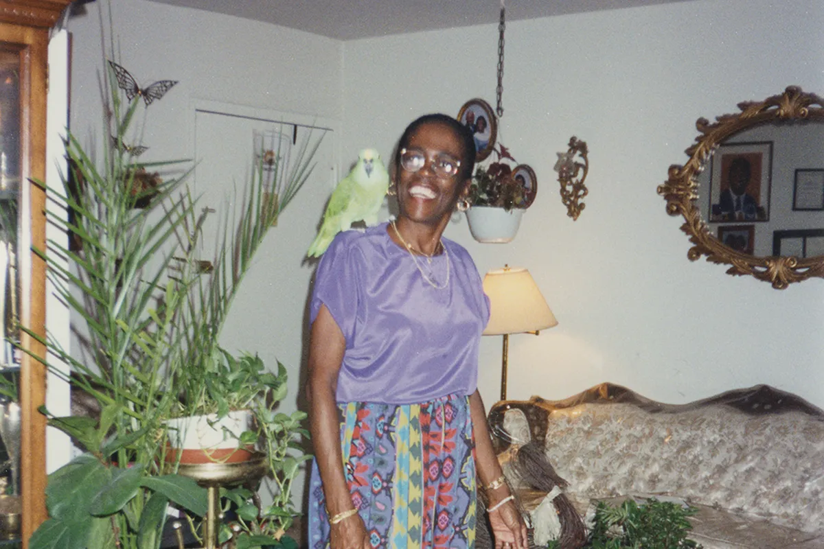 Joan Stanley standing in the middle of her living room with her green bird sitting on her right shoulder.