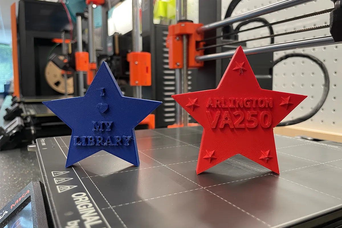 Blue and red 3D printed stars read "I heart my library" and "Arlington VA 250," with The Shop's 3D printers visible in the background.