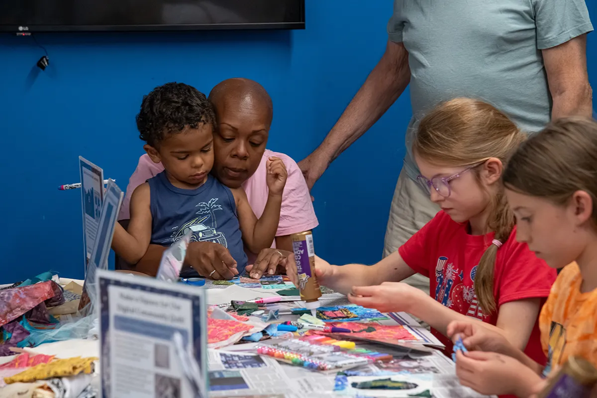 A child sits on a caregiver's lap while the pair work together to make a quilt square from fabric scraps and other supplies.