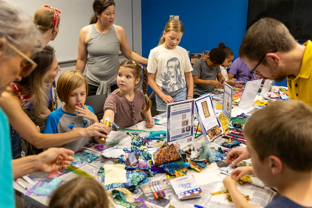 Children and families work together to make quilt squares from fabric scraps and other supplies.