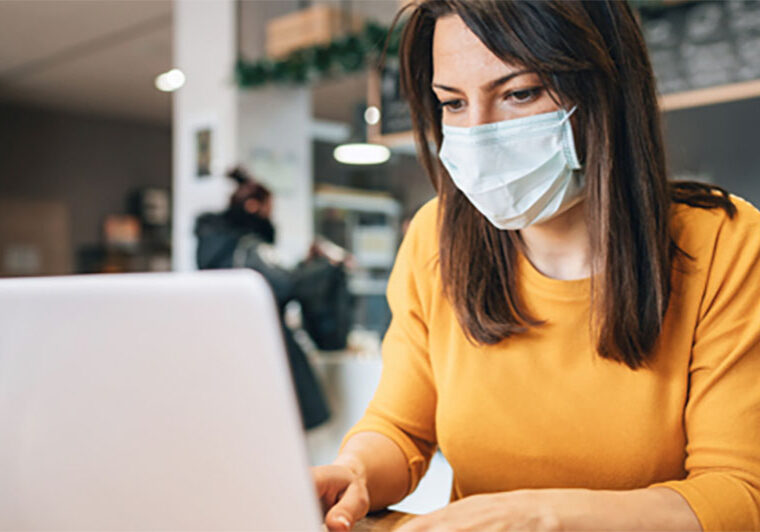Woman typing on a laptop in a coffee shop while wearing a face mask