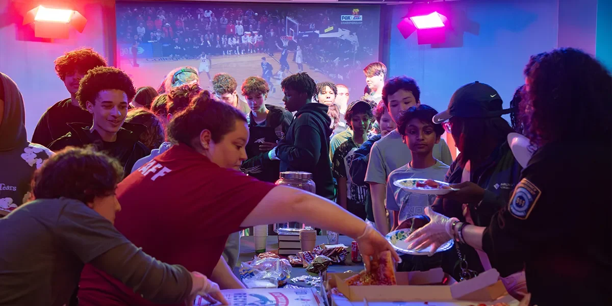 Teens crowd around a table with pizza and other food in Central Library's auditorium, with a basketball game playing on a screen in the background.