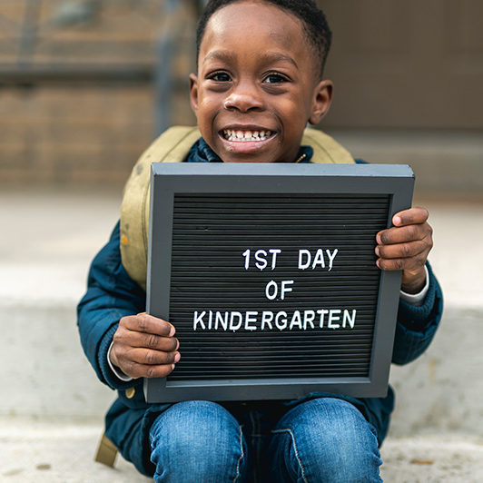 Photo of a smiling boy holding a sign depicting "1st Day of Kindergarten."