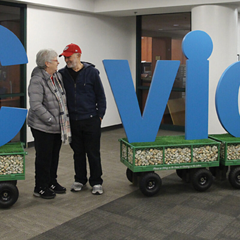 Photo of a couple standing in between a public sculture with the letters "Cvic." The couple is forming the letter "i"